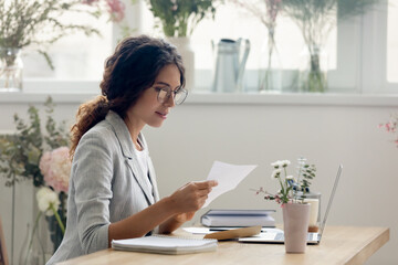 Important message. Focused attentive young woman florist designer sitting at work desk in office studio. Small business owner involved in reading paper letter from bank of credit loan terms conditions