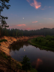 The clear evening sky and the Moon is reflected in the mirror surface of the forest river at sunset. Silence at sunset on the high bank of the forest river.