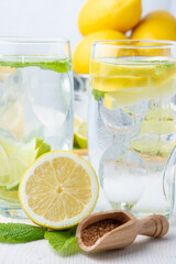 Close-up of two glasses of water with ice, brown sugar, cube with lemons and mint leaves, on white table, in vertical