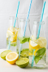 Close-up of three bottles with water, pieces of lemon and blue straws, on white wooden table with half a lemons and limes, white background, vertical
