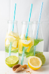 Top view of three bottles with water, lemon pieces and blue straws, on white wooden table with brown sugar, half lemons and limes, white background, vertical