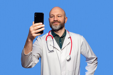 Obraz na płótnie Canvas Portrait of bald slim doctor wearing white uniform, green shirt, red stethoscope on blue background. Caucasian in 40s, grey beard, holding smart phone. Video call and online help concept