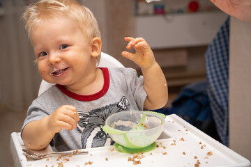 Little blond boy smiles looking at the camera because he turned over a plate of porridge and got dirty all over