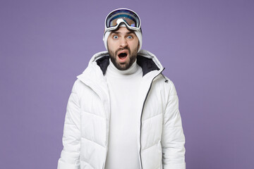 Shocked worried skier man in white windbreaker jacket ski goggles mask keep mouth open spend extreme weekend winter season in mountains isolated on purple background. People lifestyle hobby concept.