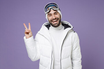 Naklejka premium Laughing skier man in white windbreaker jacket ski goggles mask showing victory sign spend extreme weekend winter season in mountains isolated on purple background. People lifestyle hobby concept.