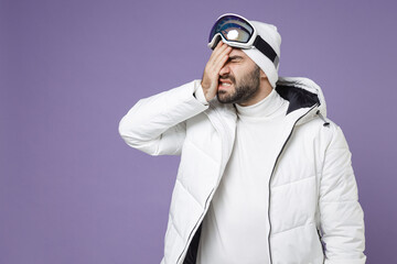Tired worried skier man in white windbreaker jacket ski goggles mask covering face with palm spend extreme weekend winter in mountains isolated on purple background. People lifestyle hobby concept.