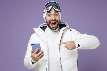Amazed skier man in white windbreaker jacket ski goggles mask point index finger on mobile cell phone spend weekend winter in mountains isolated on purple background. People lifestyle hobby concept.