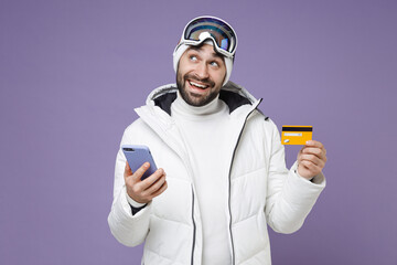 Pensive skier man in white windbreaker jacket ski goggles mask using mobile phone hold credit bank card spend weekend winter in mountains isolated on purple background. People lifestyle hobby concept.