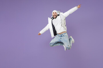 Full length of excited cheerful man in warm white padded windbreaker jacket hat jumping spreading hands isolated on purple background studio portrait. People lifestyle cold winter season concept.