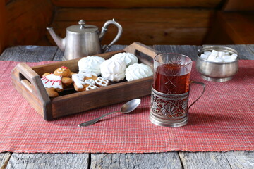 A glass of black tea is on the wooden table among silver utensils and a tray with gingerbread cookies and marshmallows.