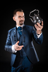 A handsome magician with a beard and mustache with a smile in a blue tailcoat holds a magic teapot in his hand