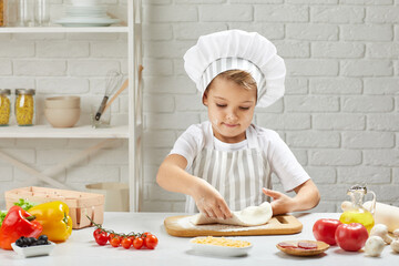 little child boy in cap and an apron knead the dough in the kitchen