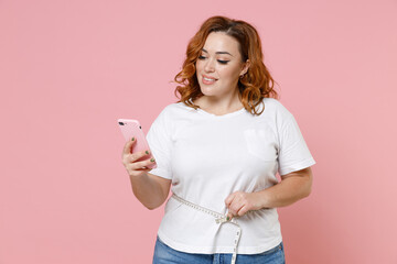 Fototapeta premium Smiling young redhead plus size body positive woman 20s wearing white casual t-shirt using mobile cell phone measuring waist with measure tape isolated on pastel pink color background studio portrait.