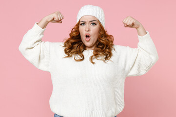 Shocked strong young redhead plus size body positive woman 20s in warm white basic sweater hat standing showing biceps muscles looking camera isolated on pastel pink color background studio portrait.