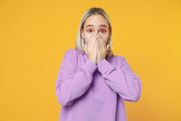 Young surprised shocked blonde caucasian woman 20s bob haircut bright makeup wearing basic purple shirt covering mouth with hands looking camera isolated on yellow color background studio portrait.