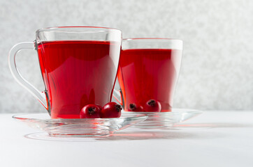 Autumn red herbal tea in two transparent cups with ripe hawthorn berries in sunlight on white wood table, closeup.