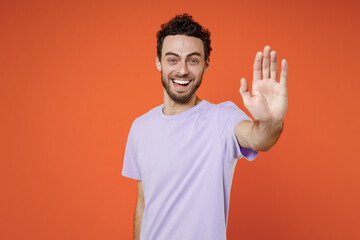Naklejka premium Cheerful laughing young bearded man 20s wearing basic casual violet t-shirt standing showing stop gesture with palm looking camera isolated on bright orange color wall background studio portrait.
