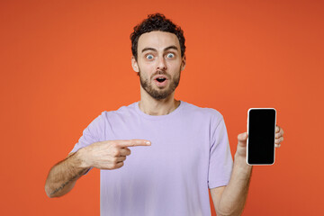 Shocked amazed young bearded man 20s in casual violet t-shirt standing pointing index finger on mobile cell phone with blank empty screen isolated on bright orange color background studio portrait.