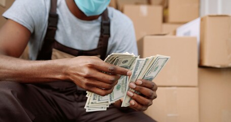 Close up of African American young handsome delivery man courier sitting in van with many boxes counting money dollars. Male postman holding money payment for shipment. Business concept