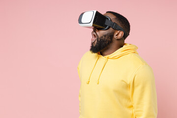 Shocked young african american man 20s in casual yellow streetwear hoodie watching in vr headset gadget rising keeping mouth open looking aside isolated on pastel pink wall background studio portrait.