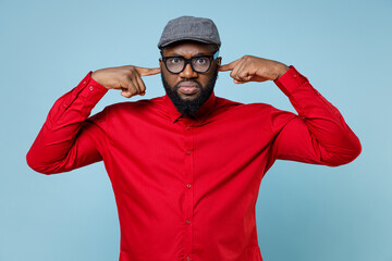 Concerned young bearded african american man 20s wearing casual red shirt eyeglasses cap standing covering ears with fingers looking camera isolated on pastel blue color background studio portrait.