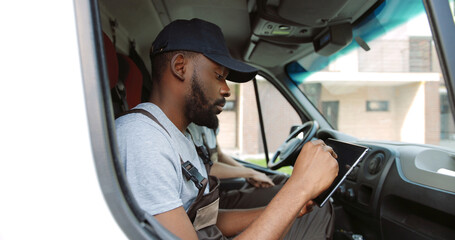 Side view of african american male courier sitting on front seat in van tapping and typing on tablet device and speaking with Caucasian colleague. Close up of delivery man in uniform using gadget