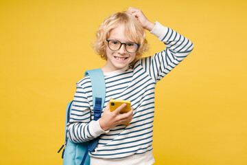 Smiling male teen boy 10s in striped sweatshirt glasses backpack using mobile cell phone typing sms message put hand on head isolated on yellow background, child studio portrait. Education concept.