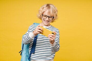 Cheerful little male kid teen boy 10s years old wearing striped sweatshirt eyeglasses backpack play game on mobile phone isolated on yellow color background, child studio portrait. Education concept.