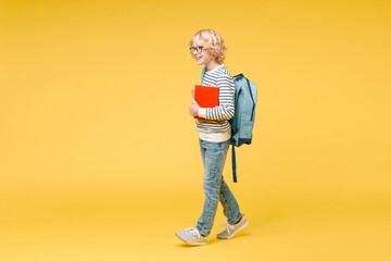 Full length side view of pretty male kid teen boy 10s in striped sweatshirt eyeglasses backpack hold school books walking going isolated on yellow background child studio portrait. Education concept.