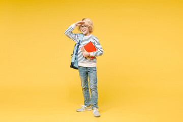 Fototapeta premium Full length of cheerful male boy 10s in sweatshirt eyeglasses backpack hold school books put hand at forehead looking far away distance isolated on yellow background child portrait. Education concept.