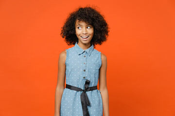 Excited little african american kid girl 12-13 years old wearing casual denim dress looking aside isolated on bright orange color background children studio portrait. Childhood lifestyle concept.
