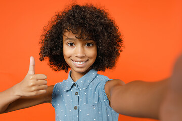 Close up of smiling little african american kid girl 12-13 in denim dress doing selfie shot on mobile phone showing thumb up isolated on orange background studio portrait. Childhood lifestyle concept.