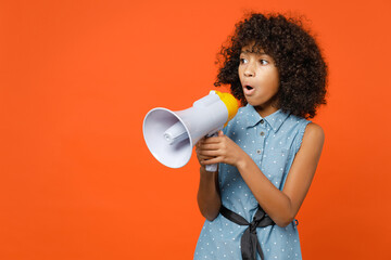 Shocked little african american kid girl 12-13 years old in casual denim dress screaming in megaphone isolated on bright orange color background children studio portrait. Childhood lifestyle concept.