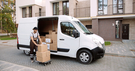 Young handsome Caucasian delivery man taking boxes from van standing on street near house delivering parcels. Male courier with ordered shopping package. E-commerce. Shipment concept
