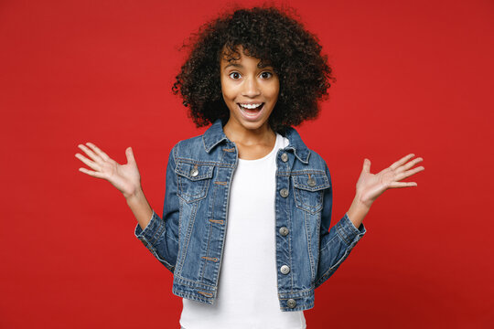 Surprised Little African American Kid Girl 12-13 Years Old Wearing Casual Denim Jacket Spreading Hands Isolated On Bright Red Color Background Children Studio Portrait. Childhood Lifestyle Concept.