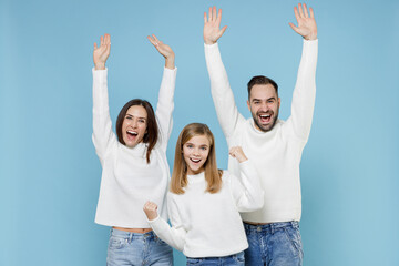 Obraz premium Happy excited young parents mom dad with child kid daughter teen girl in white sweaters doing winner gesture rising hands isolated on blue background studio portrait. Family day parenthood concept.