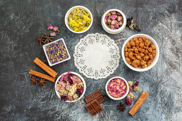 a piece of lace with bowls of dry flowers and chocolate and nut around it on grey background