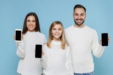 Smiling young parents mom dad with child kid daughter teen girl in white sweaters hold mobile phone with blank empty screen isolated on blue background studio. Family day parenthood childhood concept.