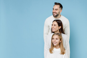 Funny young happy parents mom dad with child kid daughter teen girl in white sweaters stand behind each other looking aside isolated on blue background studio portrait. Family day parenthood concept.