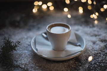 A cup of espresso surrounded by New Year's lights and pine branches in the snow. Dark background. Christmas mood