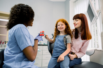 Oral care and teeth hygiene. Female African pediatric dentist explaining to young girl patient and her mother procedure of proper cleaning of teeth. Smiling child looks at camera while brushes teeth