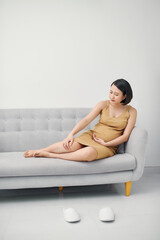 Young asian pregnant woman sitting on sofa having foot pain and leg cramps on her last trimester.