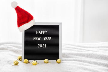 Blackboard with Santa Claus hat and text Happy New Year 2021 on white bed next to a gold Christmas balls. Home atmosphere. New Year and Christmas at home. Space for text