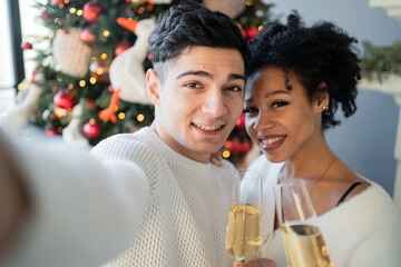 They hold champagne glasses in their hands. They are dressed in white sweatshirts. Celebrate Christmas near the Christmas tree. a couple in love take a selfie on the new year holiday.