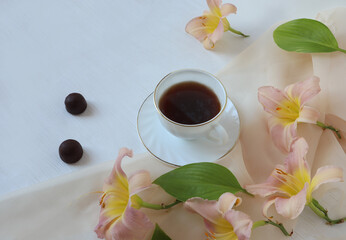 A cup of coffee with two chocolates, daylily flowers on a delicate scarf on a white background, a place for text, a top view-the concept of pleasant moments of relaxation over a cup of coffee