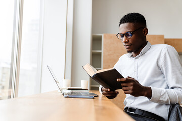 Afro american focused man reading book while working with laptop