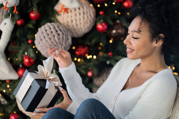 holding a surprise gift in a black box for Christmas. He wants to open it and find out what's inside. a young woman of African-American appearance is sitting on a chair near the Christmas tree.