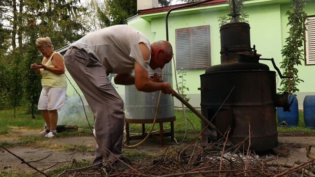 Farmer is taking out embers from fire under the distillation apparatus, for making domestic alcohol liquor, to reduce heat.