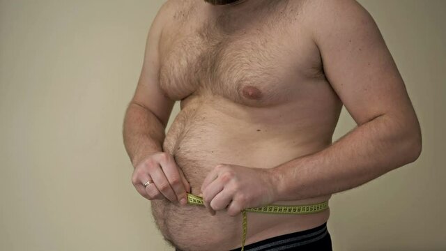 Fat man measures his waist circumference with a tape measure and shows the folds of excess fat on his stomach. Overweight problem.