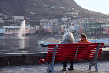 Elderly couple resting on a red bench overlooking the lake and fountain in Lugano on a warm winter day